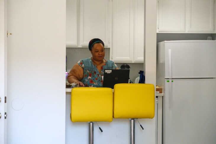 A woman wearing a blue shirt with red flowers stands in front of her laptop at the kitchen counter. There are a pair of bright yellow barstools in the foreground.