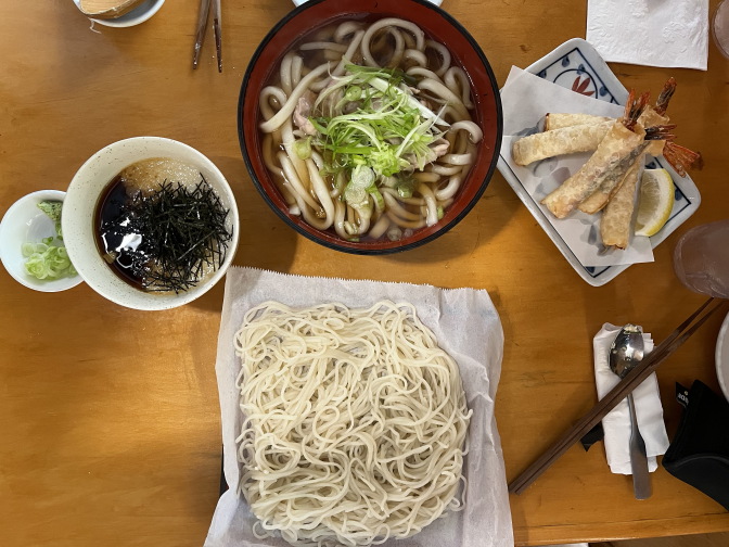 Several plates are arranged on a light-dark wooden surface. Two plates hold different types of white noodles On the right, there is a series of egg rolls with shrimp tails sticking out from the ends. To the left, a small bowl contains a cloudy white substance seasoned with a black sauce and garnished with finely shredded pieces of black seaweed known as nori.