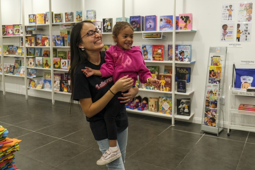 A woman with medium-light skin tone holds a 3-year-old child with medium skin tone at a children's bookstore.