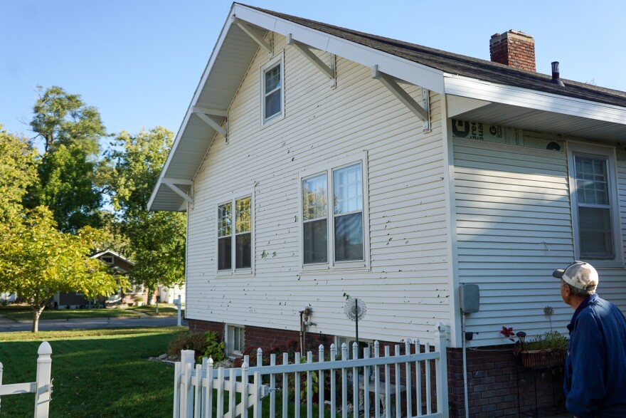 A man wearing a blue shirt and white baseball cap is pictured from behind looking at a white home. A white picket fence stands to the left of the home and there are trees in the distance