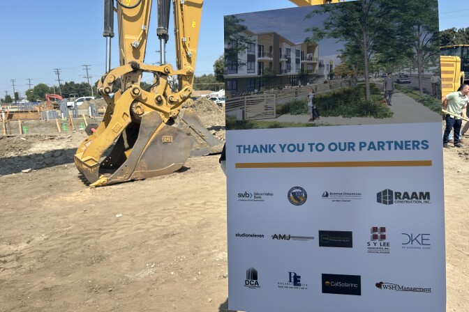 A sign in the foreground shows a rendering if an apartment complex with trees and grass out front and the words "Thank you to our partners" with company names and logos below. In the background are earth movers on a dirt lot. 