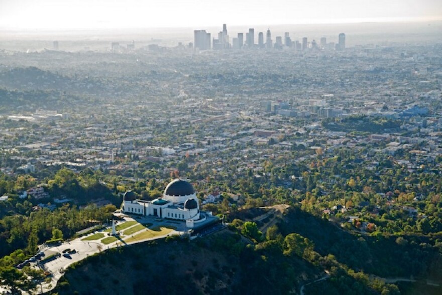 An aerial photo of the city of Los Angeles and beyond. In the foreground is the dark green capped Griffith Observatory, in the distance is the downtown L.A. skyline.