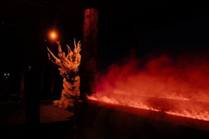 A sculpture of a tree in a dim red light to the left; on the right, steamy faux fire, the illusion of smoke burning above it.