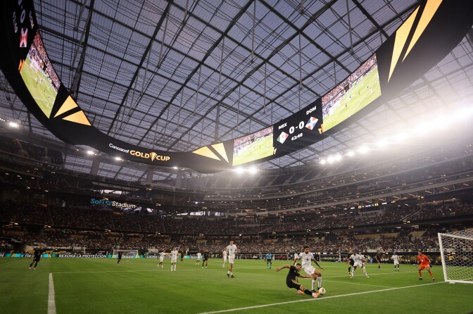 Men wearing white and black jersey's run on a green field below bright stadium lights.