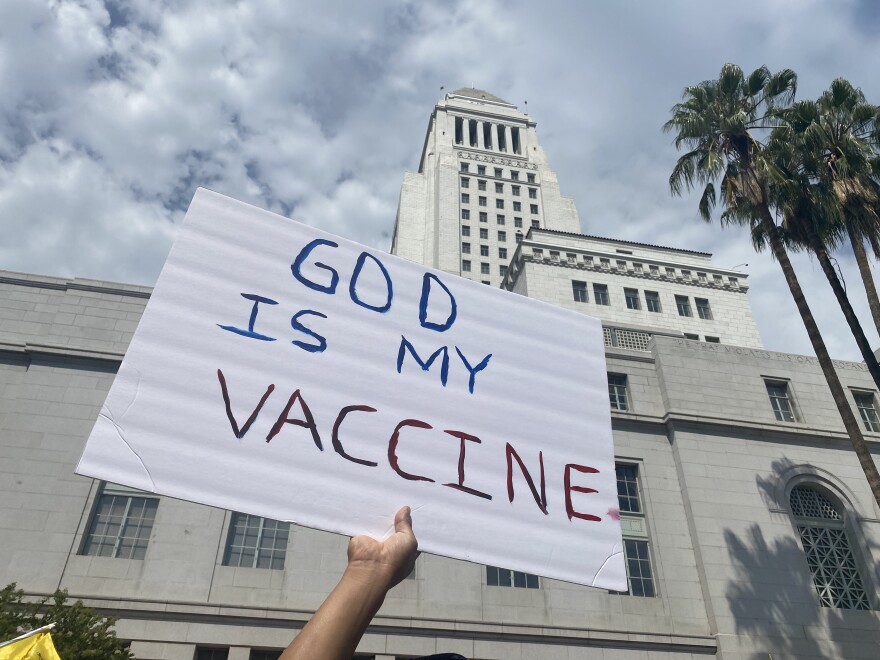 At an anti-vaccination rally in downtown LA on Aug. 14, 2021, a hand in the foreground holds up a white hand-lettered sign that says, in all capital letters, "God is my vaccine." LA City Hall towers in the background, in front of a cloudy sky.