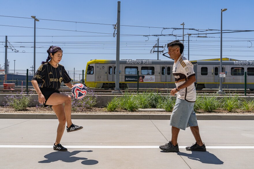 A photo of a young woman and young man playing with a soccer ball. They are standing on gray concrete next to a Metro Rail line. In the background is a blue sky and a Metro Rail train is riding by on a rail platform surrounded by fencing, light poles, and electrical wires. The woman has black hair with purple highlights and brown skin tone. She wears a black soccer jersey and black shorts and has her right knee raised to bump a soccer ball. The man wears a white soccer jersey and knee-length jean shorts. He has brown skin tone and black hair and watches the woman in anticipation of receiving the soccer ball. 
