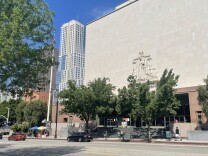 Angelenos walk to and from a a building in downtown Los Angeles. A few vehicles pass in front of the building on a treelined street, with skyscrapers in the background. 