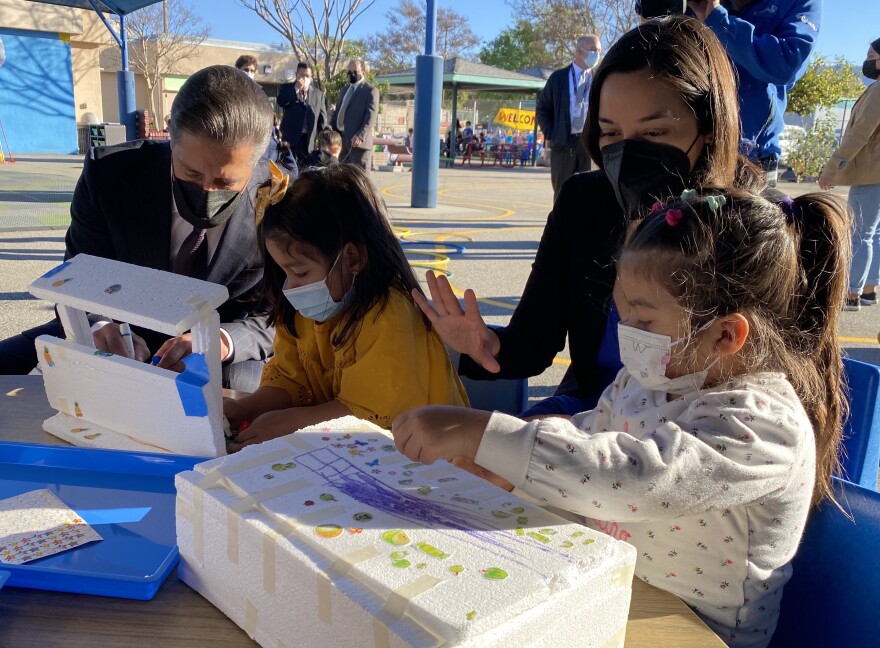 Two adults interact with two young children who are decorating and building with styrofoam blocks at an outdoor work table in a school play yard. All in the picture are wearing face coverings to prevent COVID-19 spread.