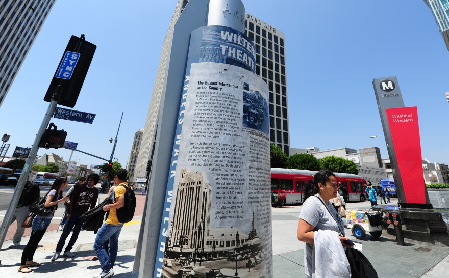 A group of four teenagers stands by a street sign by a street corner while a woman walks by a street marker that reads "Wiltern Theater." In the background is a sign for the Wilshire/Western Metro stop.