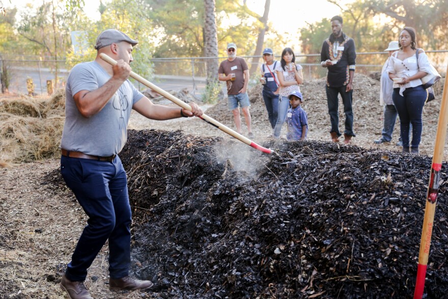 A man wearing blue pants and a grey t-shirt and cap uses a rake to dig into a large pile of compost. In the background a group of people watching. The day is sunny and trees are in the background.