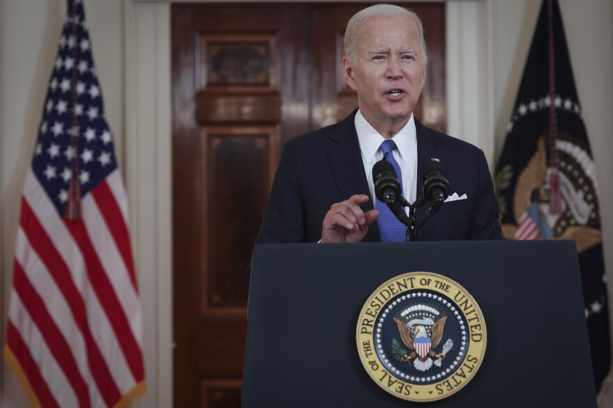 A white man with white hair speaks at a lectern with the seal of the president. He wears a dark suit with a white shirt and blue tie. A U.S. flag is over his right shoulder.