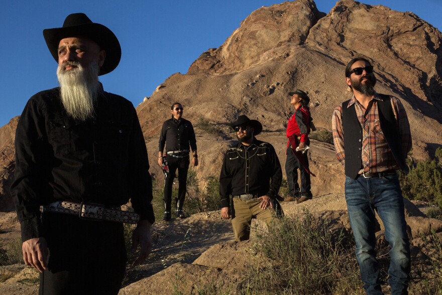 A group of five men in western-style clothing stand among rocks and shrubs in a desert landscape.