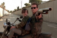 A young boy and a man wearing sunglasses and a leather jacket sit on a motorcycle, turned sideways in a flood-control channel. The man is pointing a rifle at something behind them while the boy looks at the man's face.