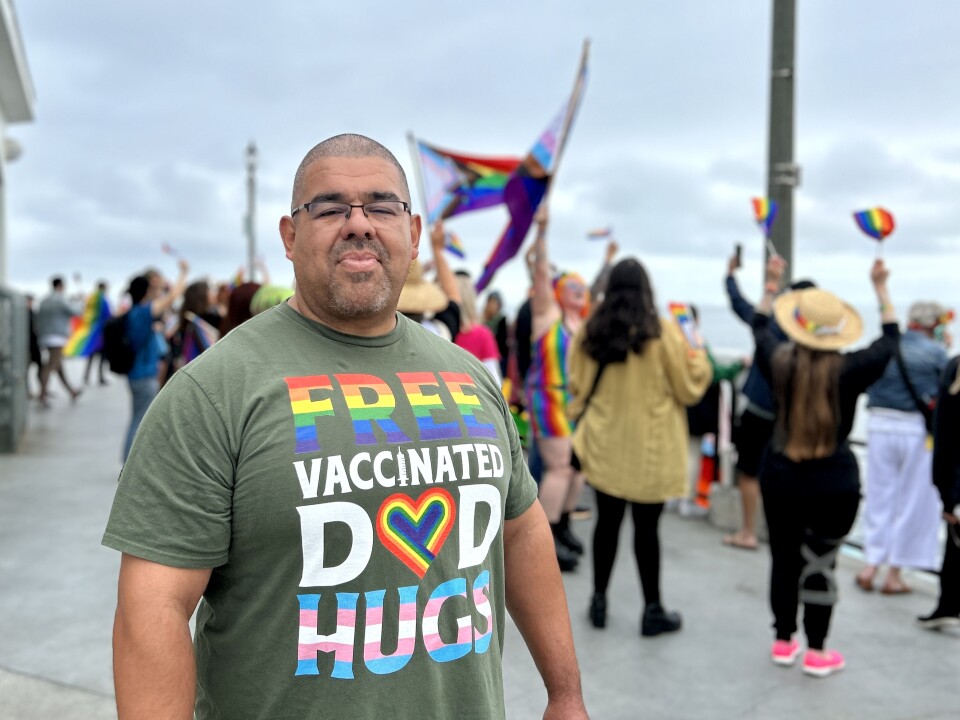 A portrait of Miguel Lopez, a man with a medium skin tone who's wearing a green shirt that says "free vaccinated dad hugs" in different Pride flag colors. In the background are people waving Pride flags.