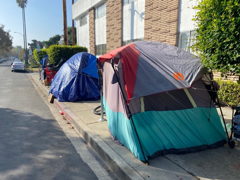 Three tents used by unhoused people are shown on a sidewalk next to an apartment building.