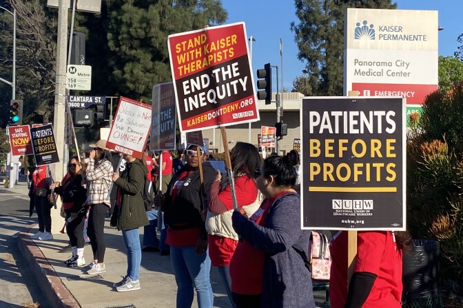 Striking mental health workers stand in line along a street in Panorama City. They hold signs that read "Patients before profits" and "End the inequity" 