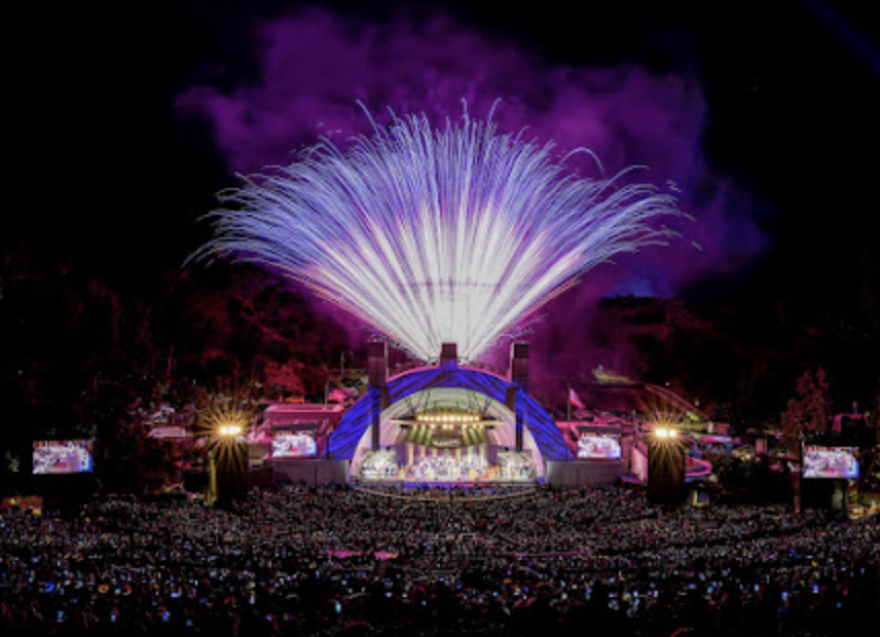 Purple fireworks light up the sky over the bandshell of the Hollywood Bowl, with the dimly lit crowd in the foregound.