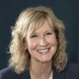 A woman with blonde, shoulder length hair, smiles while seated in front of a black background wearing a black blazer 