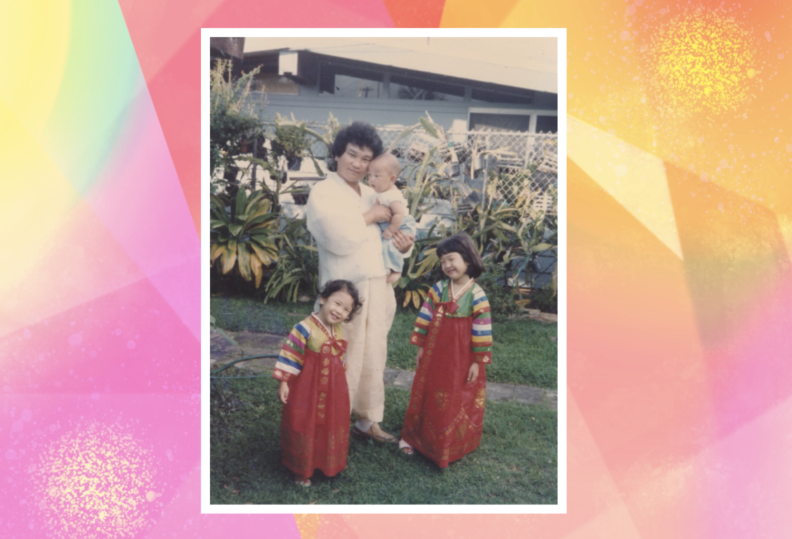 A color photo of a man in white clothing holding a baby, standing in a grassy yard. In front if him are two small children wearing red hanbok, traditional Korean dresses.