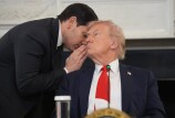 A middle-aged man in a black suit stands and whispers into the ear of an older man in a blue suit and red tie who is sitting.