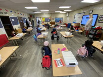 Kindergarten students sit in a classroom at desks spaced several feet apart, listening to their teacher.