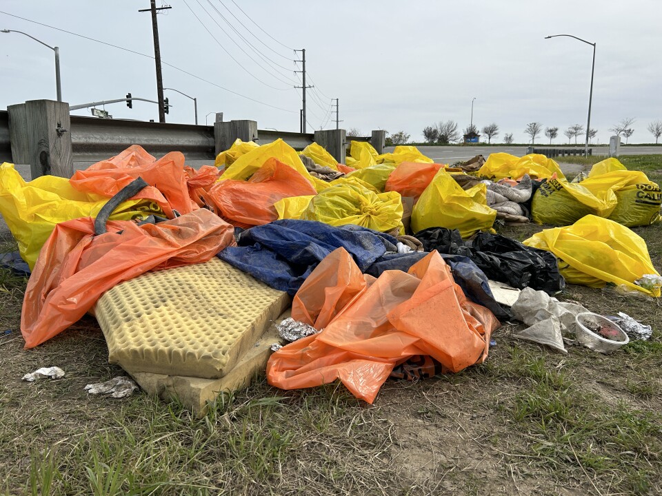 A pile of bright orange and yellow trash bags sit on a hillside next to a freeway. A dirty foam mattress poked out from underneath the pile. There are also dirty blankets and trash in the pile. 