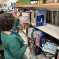 Two women stand in a library aisle in front of shelves of books. One visible title reads "Own Your Period."