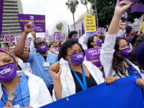 A group of women wearing purple masks with their fists in the area. There are hundreds of people around them with protests signs saying, "Residents united" and  "care for us so we can care for you."