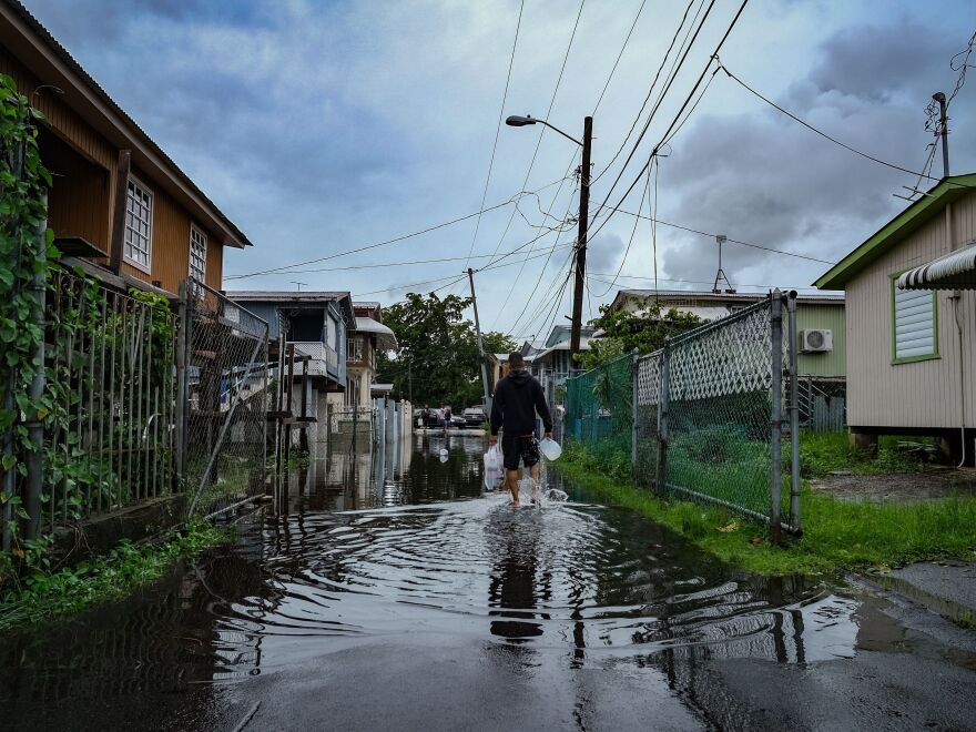A person shuffles through ankle-deep water along a flooded alley or street, gripping a plastic jug in each hand.