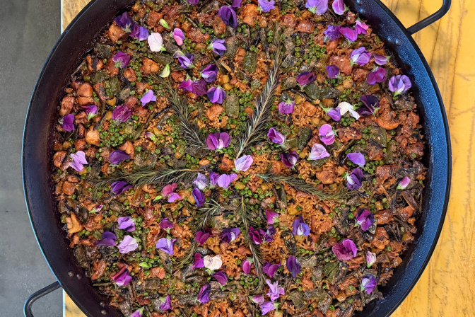 An overhead shot of a large black paella pan filled with saffron-orange rice studded with chicken, charred green vegetables, and green peas, arranged in a starburst pattern with sprigs of rosemary and scattered purple and pink flower petals. The pan sits on a mustard-yellow tabletop.