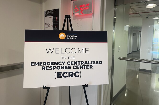 A white sign with a black stripe across the top that says "Homeless Initiative". The sign says "Welcome to the Emergency Centralized Response Center (ECRC). The sign is in the entrance to an office building.