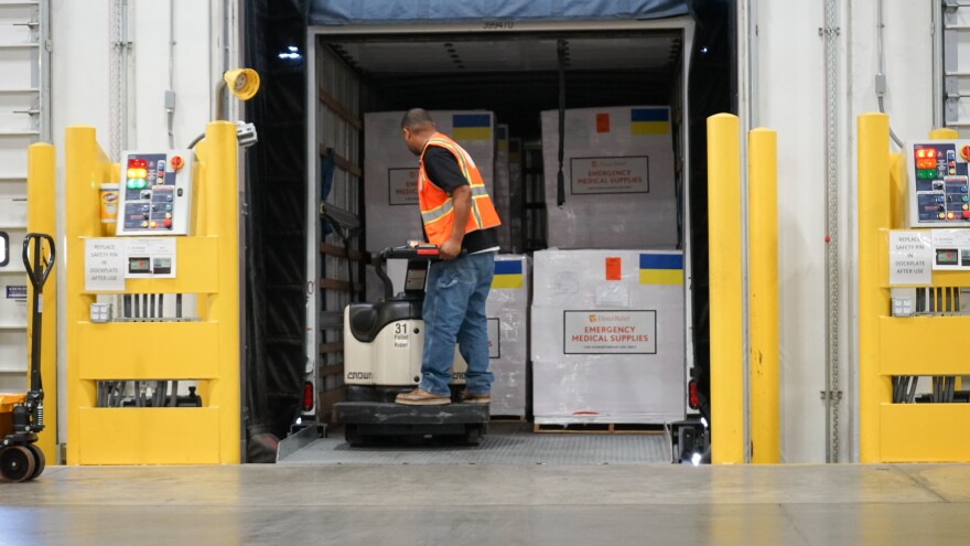 A man wearing an orange vest stands in the back of a truck in a loading bay. He's surrounded by large metal boxes labeled "Emergency Medical Supplies,", each with a Ukrainian yellow and blue flag sticker.