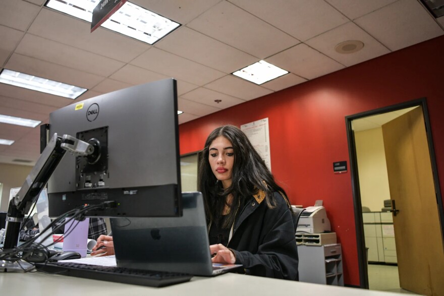 A young woman with fair skin and long dark hair sits in front of a laptop and desktop monitor. She wears a baggy black coat and looks at her laptop screen.