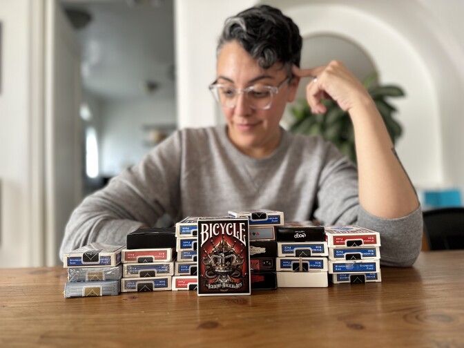 A person with medium-toned light skin and short bobbed haircut, wearing eyeglasses and a gray sweatshirt, looks at a stack of more than 20 packs of cards on their living room table. 