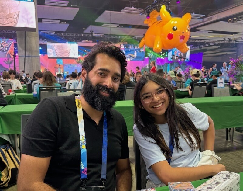 A man with medium skin tone and a bushy dark beard sits next to a girl with medium skin tone and long dark hair. Behind them are several tables filled with people playing a card game, above whom floats a large yellow Pikachu.