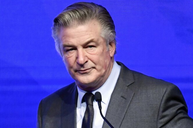 A close up shot of a clean cut, male presenting person in a dark suit speaking into a microphone in front of a blue background