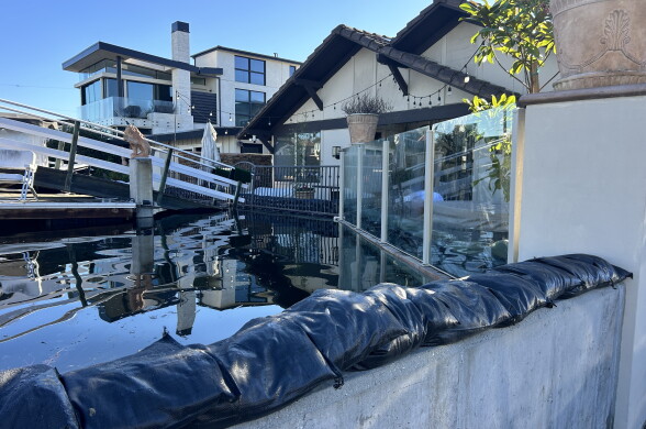 Sandbags line a wall as water rises near homes and a dock in a waterfront neighborhood.