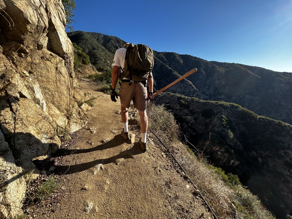 A wide shot of the back of an older white man hiking up a rocky dirt trail with mountains in the background on a sunny day. 