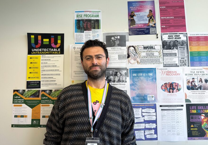 A man with dark hair, short beard and glasses is seen standing against a wall of flyers.