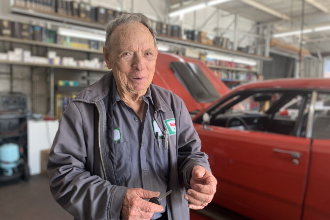 An older man in an auto repair shop uniform stands in a body shop with a red car behind him.
