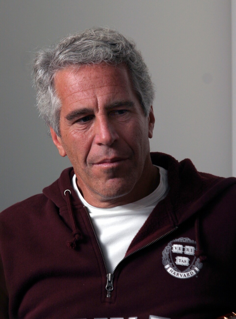 A man with white hair wearing a burgundy sweater with a zipper and a white tshirt underneath looks to his left, expressionless
