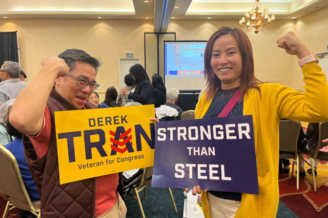 An Asian man holds a sign that reads "Derek Tran/ Veteran for Congress" while holding an arm up in a show of strength stands next to an Asian woman making the same gesture while holding a sign that reads "Stronger than Steel."