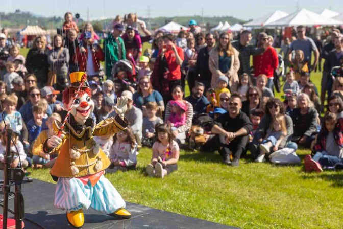 A crowd of hildren and their parents are on a grass field watching marionnette show in the foreground. 