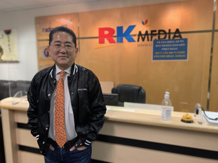 A Korean American man poses with a faint smile in the lobby of a radio station. He is wearing glasses, with a dark jacket that has the Radio Korea insignia embroidered on the left side, over a white shirt and patterned tie. Lettering that reads "RK Media" is on the wood wall paneling behind him.