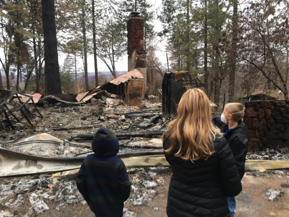 A woman with long reddish blond hair stands with two children in front of charred debris at the site of a wildfire