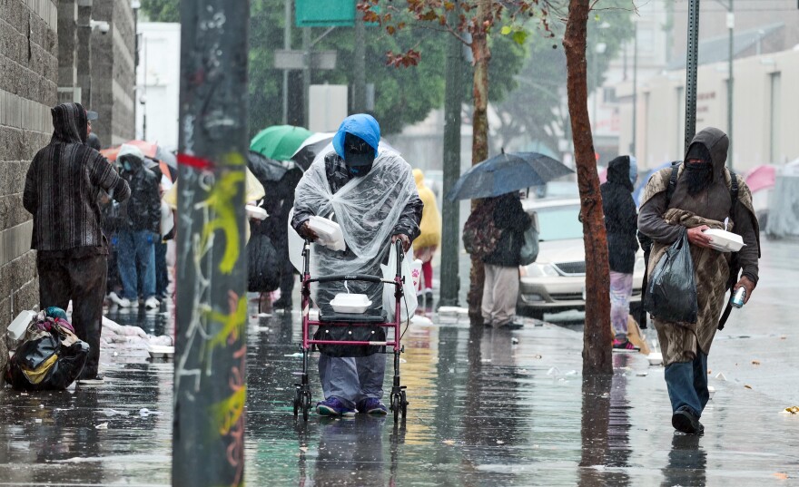A busy city street in the middle of a rain storm. Several people are standing on the sidewalk with umbrellas and coats while a person wearing a plastic poncho pushes a mobility scooter towards the camera.