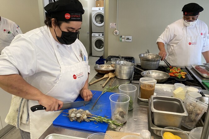 Culinary apprentice Nicole Rios, in a white chef's outfit with a black facemask, stands in a kitchen cutting a piece of rosemary on a blue cutting board next to a pile of shrimp skewered on rosemary twigs while another chef grills vegetables nearby.