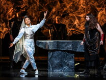 A woman in a veil and a sparkling pantsuit is joined by two men dressed as Greek gods on a stage around a stone table.