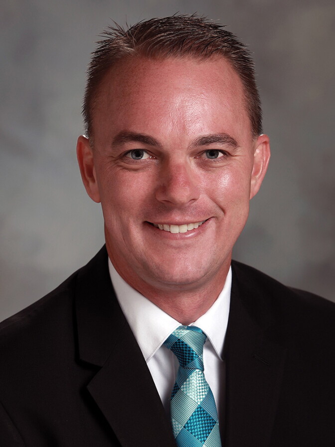 A headshot of a male presenting person with a light skin tone in a black suit and teal tie.