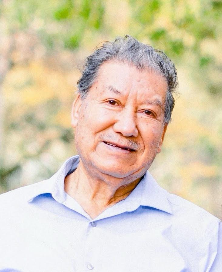 A man with medium skin tone with some facial hair, gray hair, wearing a light blue button down shirt, smiles at the camera.
